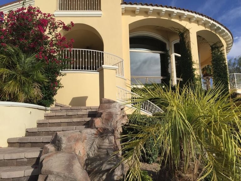 Curved yellow Mediterranean-style villa with stone steps, arched features, and tropical plants including pink bougainvillea and palm trees